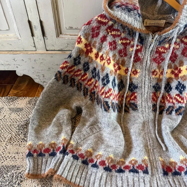 A cozy knit zip-up hoodie sweater with a gray heather base and colorful Fair Isle patterns in red, yellow, navy, and brown, laid flat on a patterned rug in front of a distressed white cabinet.