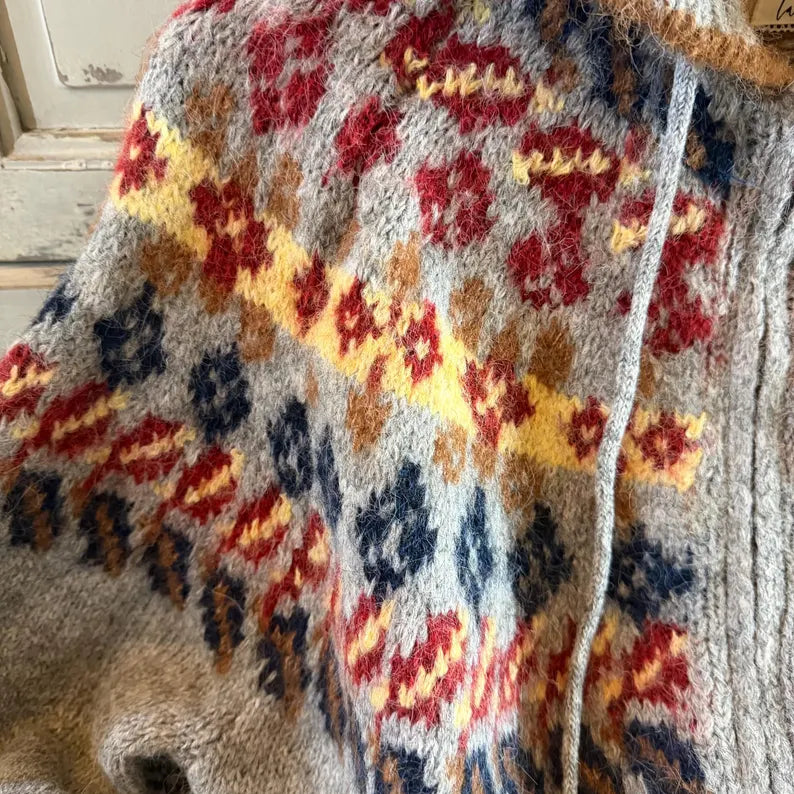 A close-up of a fuzzy knit sweater or poncho with a multicolor Fair Isle pattern on a light gray base, featuring red, yellow, navy, and brown motifs.