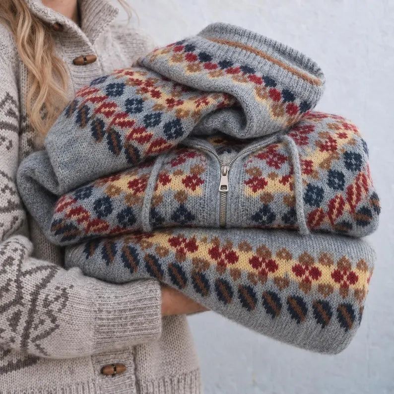 A person holds a folded gray knit zip-up sweater with a multicolor Fair Isle pattern in red, blue, yellow, and brown. The sweater has a drawstring hood and ribbed cuffs and hem.