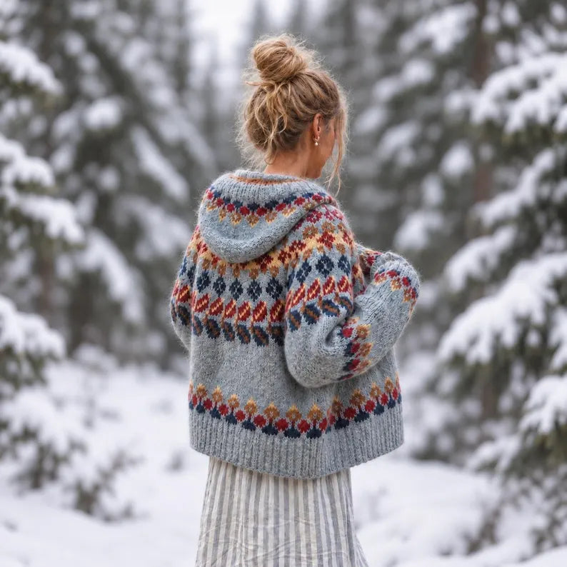 A woman stands with her back to the camera in a snowy forest, wearing a light gray knitted hooded sweater with a colorful Fair Isle pattern in red, orange, blue, and yellow.