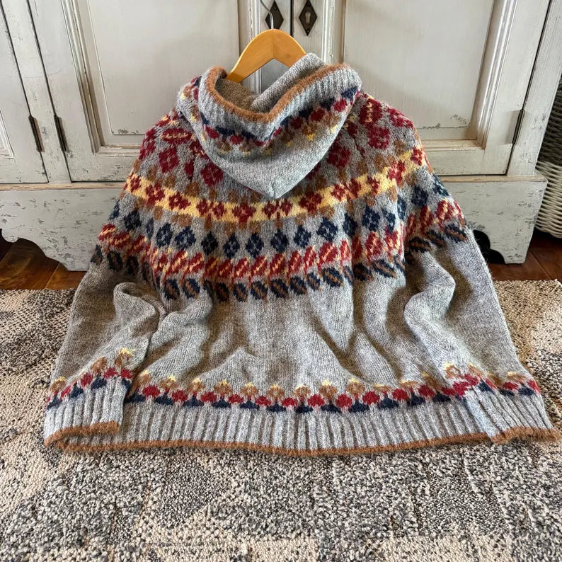 A chunky knit cowl-neck sweater in heather gray with a multicolor Fair Isle pattern, hanging on a wooden hanger on a textured rug in front of a white cabinet.