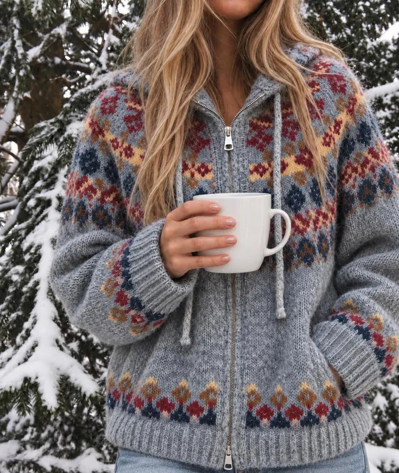 A woman in a snowy outdoor setting wearing a cozy gray zip-up hoodie with a multicolor Fair Isle pattern. She is holding a white ceramic mug.