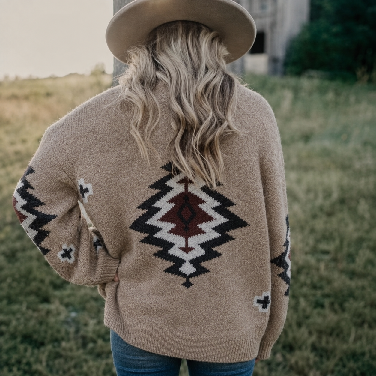 A woman viewed from behind wearing a beige knit sweater with a Southwestern geometric pattern in black, white, and dark red. She is also wearing a wide-brimmed beige hat and blue jeans, standing outdoors in a grassy field.