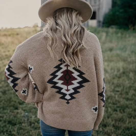 A woman viewed from behind wearing a beige knit sweater with a Southwestern geometric pattern in black, white, and dark red. She is also wearing a wide-brimmed beige hat and blue jeans, standing outdoors in a grassy field.