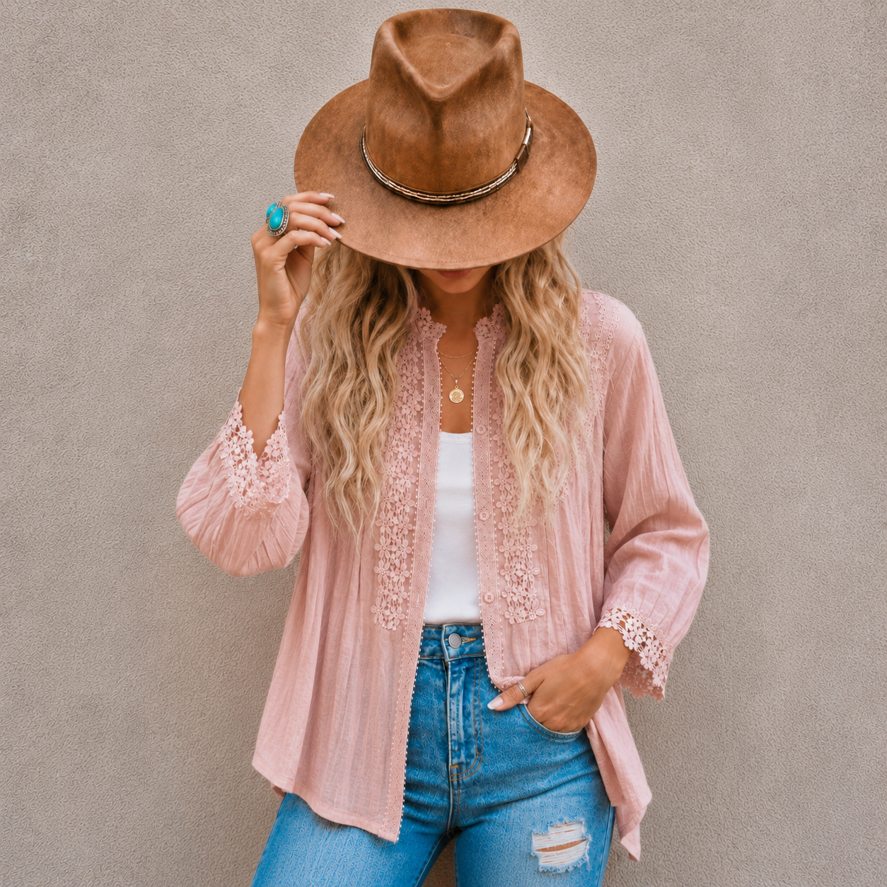 A woman wearing a light pink lace-trimmed cardigan, a brown wide-brim hat, a white tank top, and blue jeans, posing against a textured gray wall.