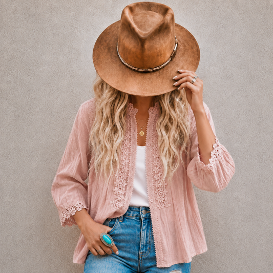 A woman wearing a light pink lace-trimmed button-up shirt, a brown wide-brim hat, a white top, and blue jeans, standing against a textured gray wall.