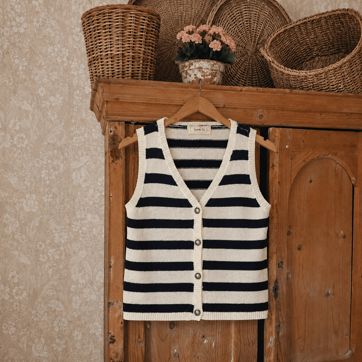 A sleeveless knit cardigan vest with horizontal navy and cream stripes, hanging on a wooden hanger from a rustic wooden cabinet. The cabinet is decorated with woven baskets and a small potted plant against a textured beige wallpaper background.