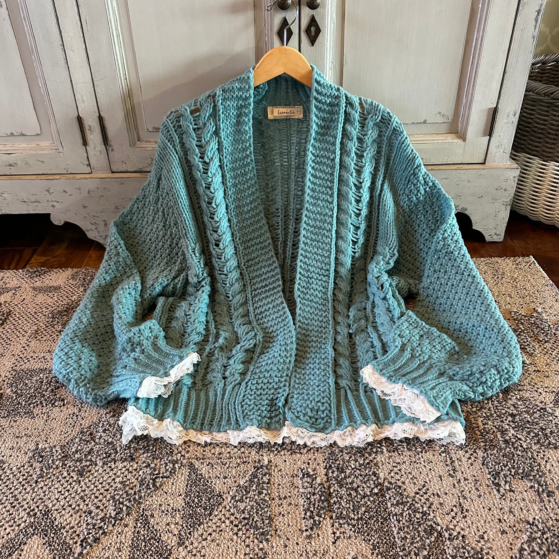Teal knitted cardigan with lace trim on a wooden hanger against a wooden cabinet background.