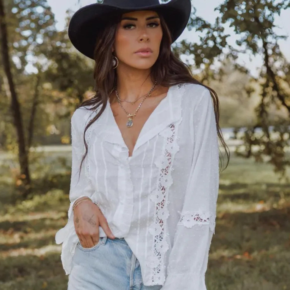 Woman wearing a white blouse with lace details and a black hat in a natural setting.