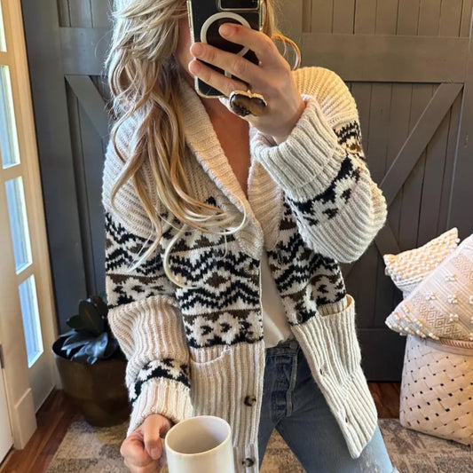 Person wearing a patterned cardigan holding a phone and a mug indoors.