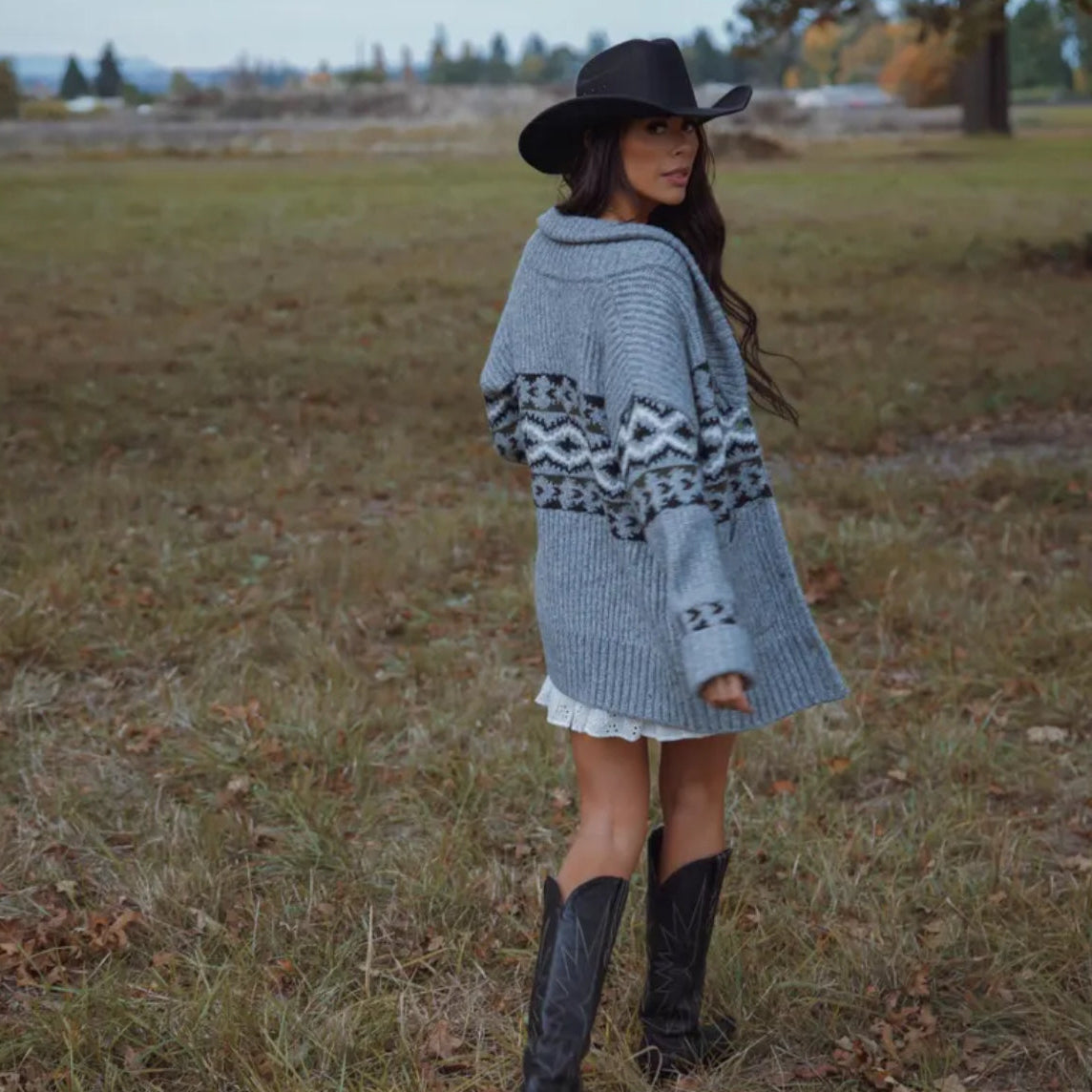 Woman wearing a gray patterned sweater, black cowboy hat, and boots in an open field.