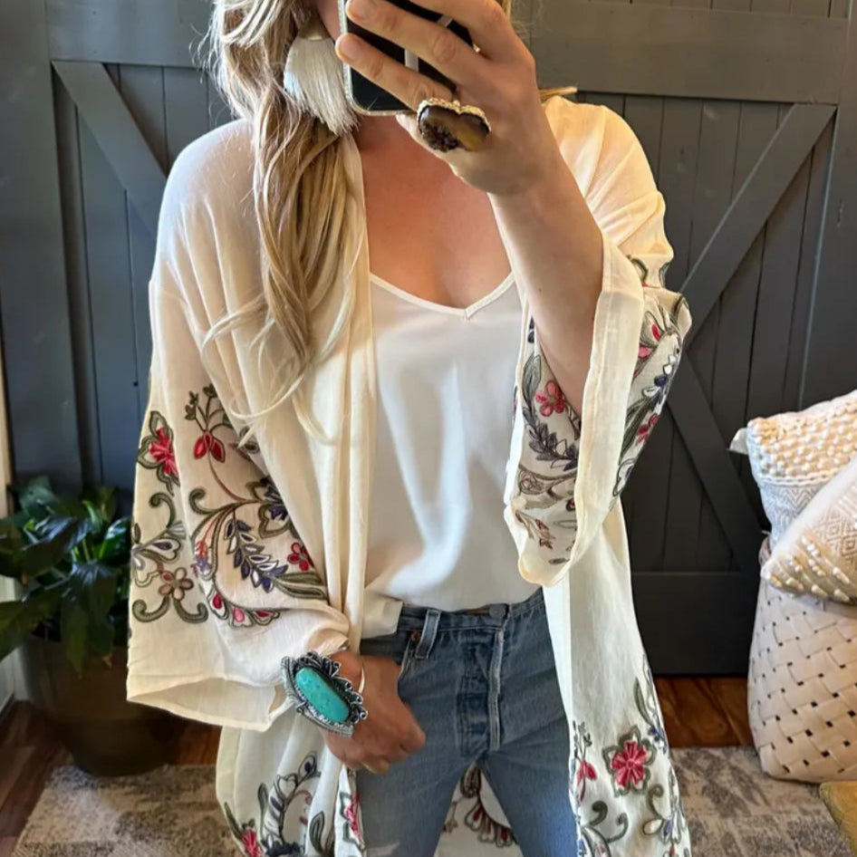 Person wearing a floral embroidered cardigan over a white top and jeans, standing in a room with wooden walls and a plant.