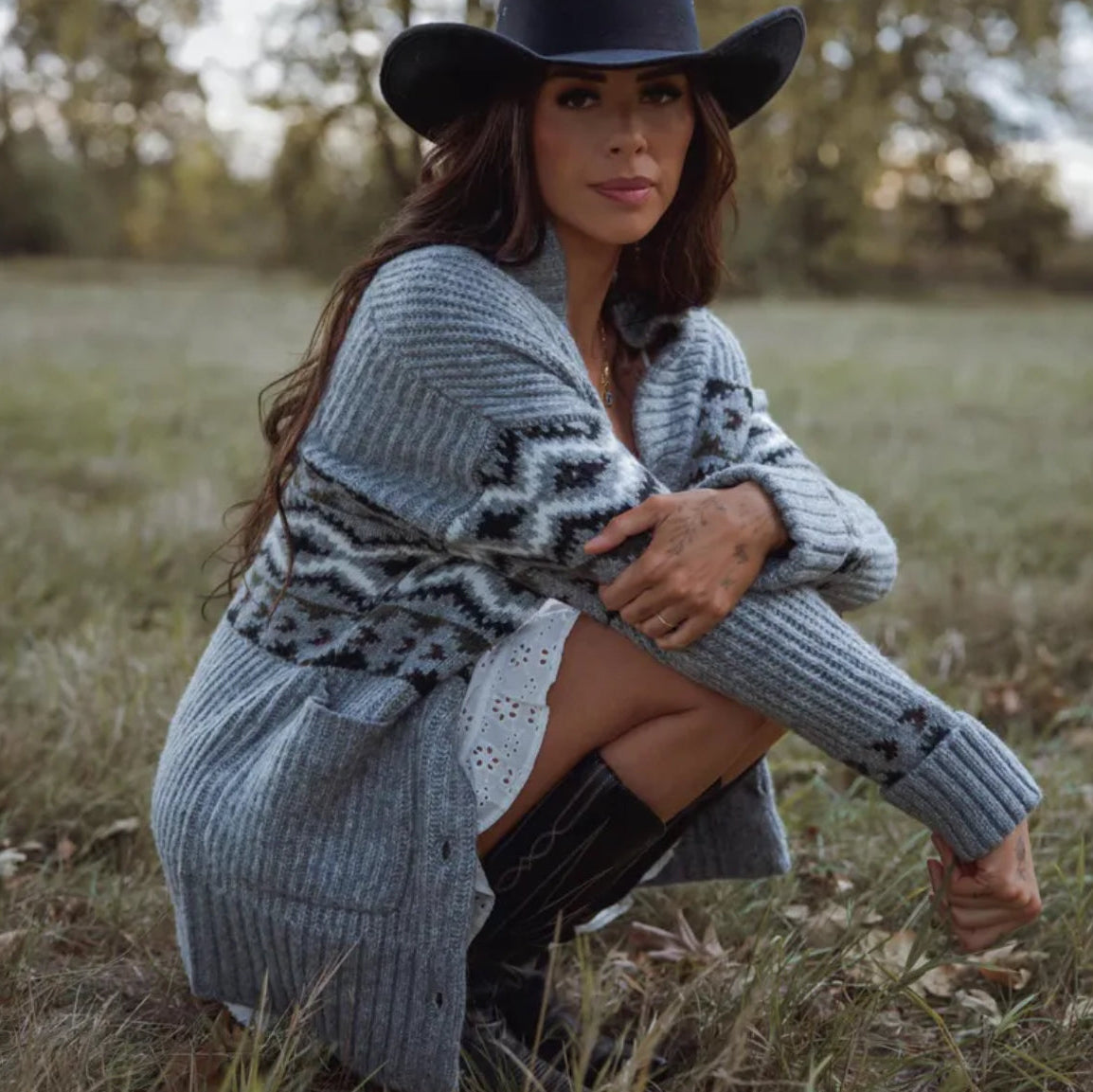 Woman in a patterned cardigan and wide-brimmed hat sitting in a field.