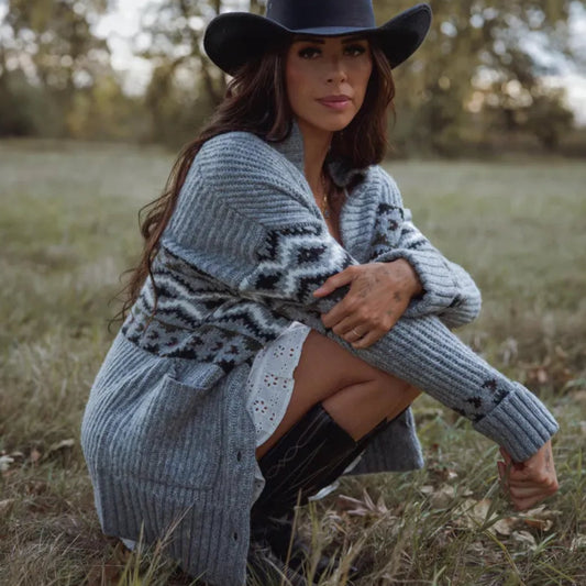 Woman in a patterned cardigan and wide-brimmed hat sitting in a field.