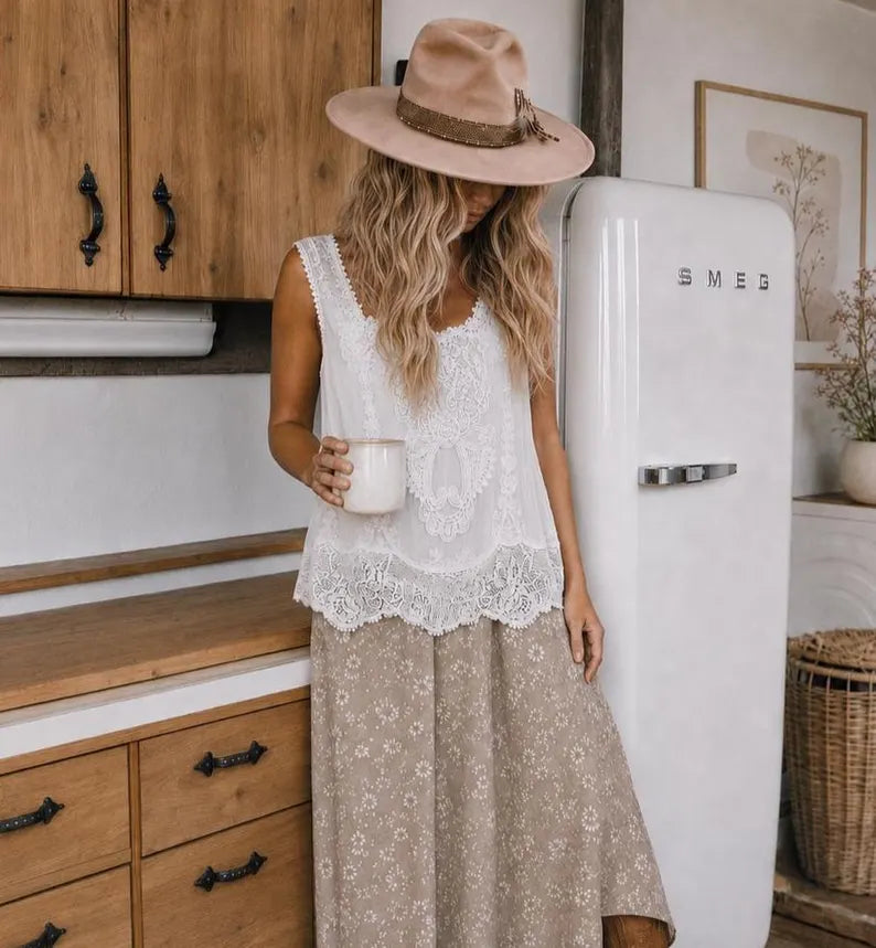 A woman stands in a rustic kitchen wearing a white lace sleeveless top, a beige wide-brim hat, and a beige floral skirt, holding a white mug next to a vintage white SMEG refrigerator.