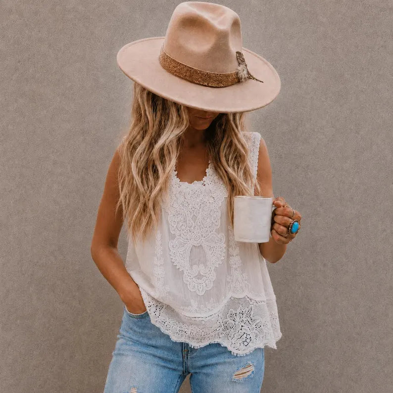 A woman wearing a beige wide-brim hat, a white lace sleeveless top, and light blue jeans, holding a white mug against a textured gray wall.