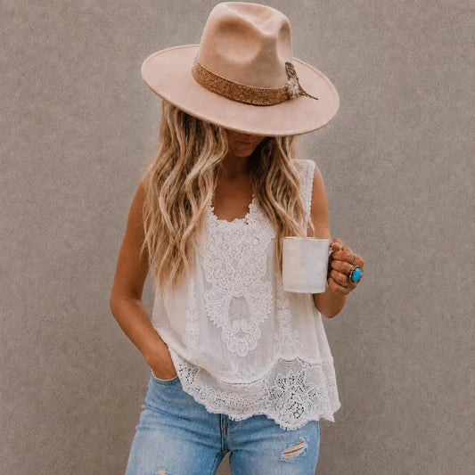 A woman wearing a beige wide-brim hat, a white lace sleeveless top, and light blue jeans, holding a white mug against a textured gray wall.
