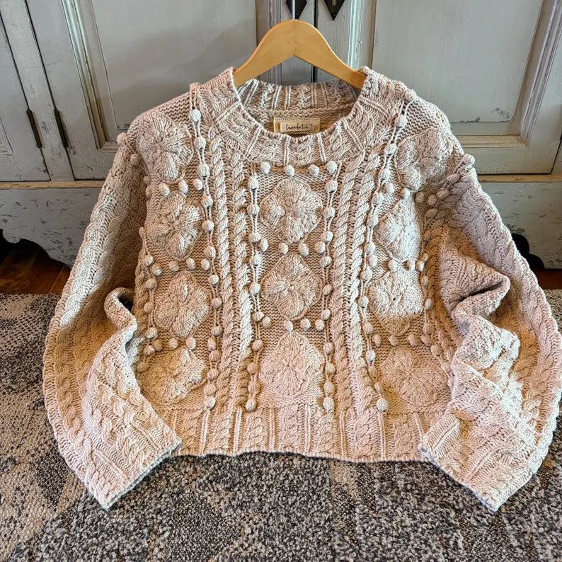 A cream-colored cable-knit sweater with textured pom-pom details, displayed on a wooden hanger over a patterned rug in front of a rustic white cabinet.