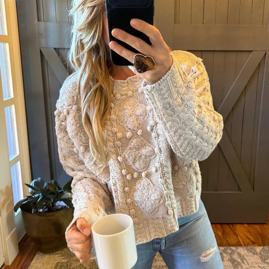 A woman takes a mirror selfie wearing a cream-colored cable-knit sweater with pom-pom details, paired with light-wash blue jeans. She is holding a white ceramic mug in a home setting with a wooden barn door background.