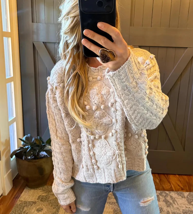 A woman takes a mirror selfie wearing a cream-colored cable-knit sweater with pom-pom details, paired with light-wash distressed jeans. She is standing in front of a dark wood barn-style door and a potted plant.