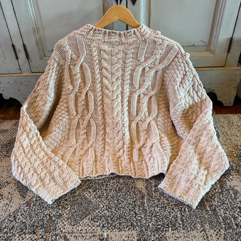 A cream-colored cable-knit cropped sweater with long sleeves, displayed on a wooden hanger over a patterned rug in front of a rustic white cabinet.