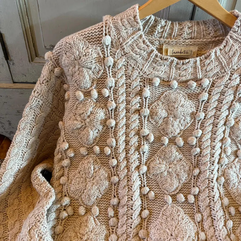 A close-up of a cream-colored chunky knit sweater with textured floral patterns, vertical cable knit details, and pom-pom accents, hanging on a wooden hanger.