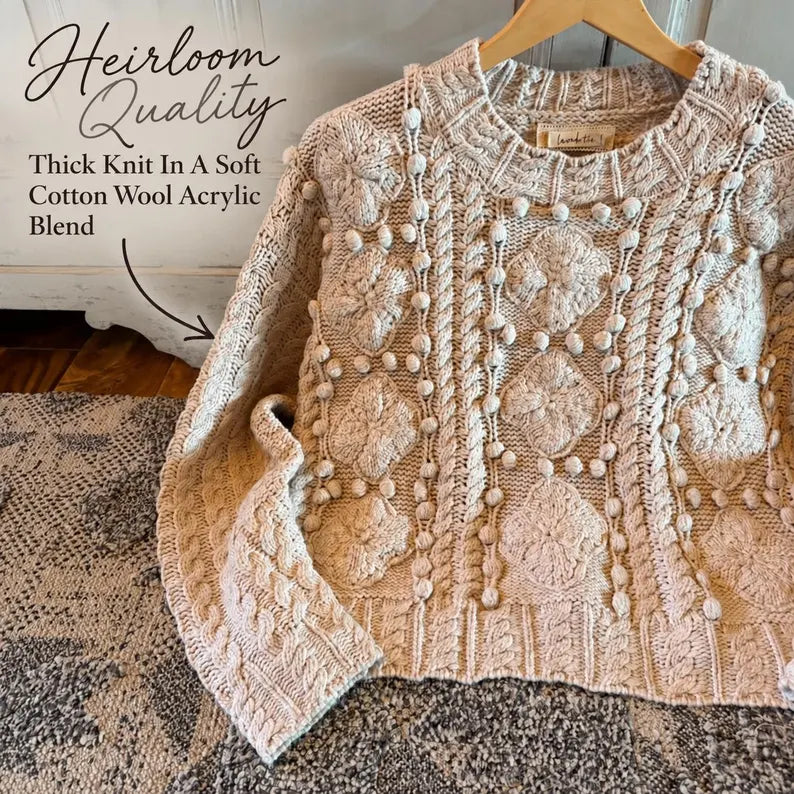 A cream-colored cable-knit sweater with pom-pom detailing, displayed on a wooden hanger on a textured rug. Text overlays on the top left describe the 'Heirloom Quality' thick knit in a soft cotton wool acrylic blend.