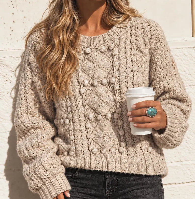 A woman wearing a chunky cable-knit sweater in a warm beige/taupe color, holding a white coffee cup against a light textured wall.