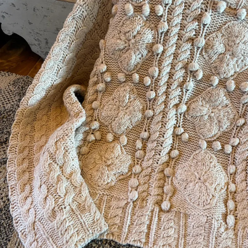A close-up of a chunky knit blanket or throw in a warm cream or light beige color, featuring intricate cable-knit patterns, raised floral motifs, and rows of small pom-poms.